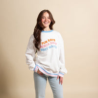 A smiling woman stands against a beige background wearing The Darling Effect’s Sun Rays, Beach Days, Boat Waves Oversized Crew—an oversized white sweatshirt with pastel trim and embroidered lettering, perfect for a cozy wardrobe.