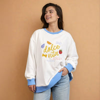 A woman smiles to the side against a tan background, wearing The Darling Effect’s Dolce Vita Oversized Crew—an ultra-cozy white sweatshirt featuring embroidered Italian icons and dolce vita ITALIA text.