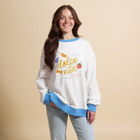 A smiling woman with long brown hair wears The Darling Effect's Dolce Vita Oversized Crew, a white sweatshirt with blue cuffs and colorful embroidery, styled with light blue jeans against a beige background.