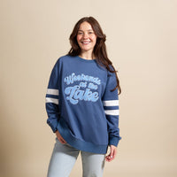A smiling woman wears The Darling Effect’s Weekends at the Lake Oversized Crew, a blue sweatshirt with chenille lettering and white sleeve stripes, styled effortlessly with light wash jeans.