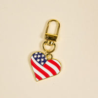The Darling Effect's Americana Heart Pawfect Collar Charm is a gold keychain featuring a heart-shaped American flag design—perfect for adding patriotic flair and unique style to your pet’s collar.