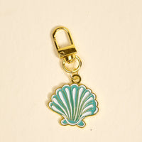 The Darling Effect’s Seashell Pawfect Collar Charm is a gold keychain featuring a blue and white enamel seashell, perfect for adding chic style to your keys or your pet’s collar on a plain light background.