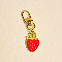 The Darling Effect's Strawberry Pawfect Collar Charm features a gold keychain with a red strawberry and green leaves, perfect as a playful charm for pet collars, shown on a cream-colored background.