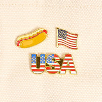 USA The Pin Pack by The Darling Effect features three bold pins—a hot dog, an American flag, and a "USA" design with stars and stripes—on beige fabric for a standout red, white, and blue set.