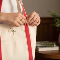 A person adjusts the strap of a cream-colored tote with red handles, decorated with The Darling Effect’s Vacation Mode The Pin Pack—pins featuring a drink, sunglasses, palm tree, and seashell. Books and a green plant sit on a table in the background.