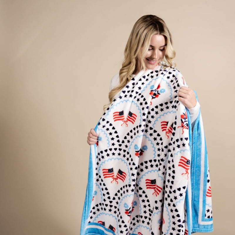A smiling woman with long blonde hair holds The Darling Effect’s All-American Darling Snuggle Up Plush Blanket, featuring American flags, blue stars, and eagles with a blue border, against a beige background.
