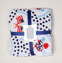 The Darling Effect’s All-American Darling Snuggle Up Plush Blanket features white plush fabric with blue, red, and navy stars, hearts, flowers, and American flags—finished with a blue ribbon. Cozy up anytime with this super-soft blanket.