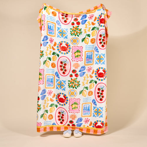 A person stands behind The Darling Effect's Ciao Bella Snuggle Up Plush Blanket, featuring a playful fruit and floral design. Only their white sock-clad feet peek out from under the large, colorful blanket against a plain beige background.
