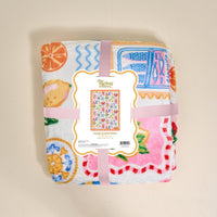 A folded, colorful Ciao Bella Snuggle Up Plush Blanket by The Darling Effect features fruit and kitchenware prints, wrapped in a pink ribbon. The label displays the brand name and a small image of the blanket’s full design.