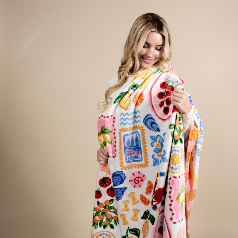 A smiling woman holds The Darling Effect's Ciao Bella Snuggle Up Plush Blanket, featuring playful fruit, seafood, and postage stamp designs.