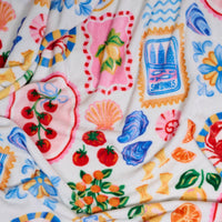 The Darling Effect’s Ciao Bella Snuggle Up Plush Blanket features bright illustrations of tomatoes, oranges, sardines, lemons, seashells, and flowers in vibrant red, blue, yellow, orange, and pink on a soft white background.