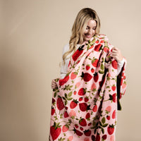 A smiling woman with long blonde hair wraps herself in The Darling Effect's Strawberry Fields Snuggle Up Plush Blanket, a soft pink throw with red strawberries and green leaves, standing against a plain beige background.