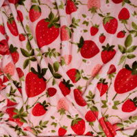The Darling Effect’s Strawberry Fields Snuggle Up Plush Blanket features a pink background with red and pink strawberries and green leaves, offering a soft, beautifully draped, slightly wrinkled plush fabric.