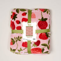 The Darling Effect's Strawberry Fields Snuggle Up Plush Blanket is a folded pink throw with red strawberries, tied with a green ribbon and a matching strawberry-themed label.