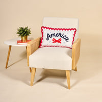 A modern cream armchair with wooden arms features The Darling Effect’s America Scalloped Rectangle Throw Pillow. Beside the chair, a white side table holds a potted plant and two stacked books.