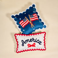 The Darling Effect's America Scalloped Rectangle Throw Pillow set features one blue pillow with embroidered flags and stars, and a white pillow with red trim, "America" text, a red bow, and 1776—perfect accent pieces.
