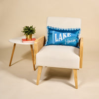 A white armchair with wooden legs and arms features The Darling Effect’s Lake House Scalloped Rectangle Throw Pillow in blue. Beside it, a white side table holds a potted plant and two stacked books against a beige background.
