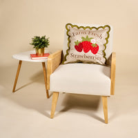 A cream armchair featuring The Darling Effect’s Farm Fresh Strawberries Scalloped Square Throw Pillow and a cozy layer sits beside a small white table topped with a potted plant.