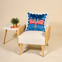 A modern white armchair with wooden accents holds The Darling Effect’s American Flags Scalloped Square Throw Pillow. Beside it sits a small white table topped with a potted plant and two stacked books.