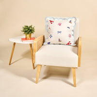 A white armchair with wooden legs holds The Darling Effect’s Seaside Haven Scalloped Square Throw Pillow with nautical designs. Next to it, a small white table with wooden legs features a potted plant and two stacked books against a beige background.