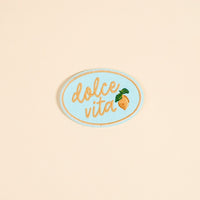 The Darling Effect's Dolce Vita Mix & Match Iron-On Patch features an oval shape, light blue background, gold cursive "dolce vita," and a yellow lemon with green leaves—perfect for brightening up your patch collection.