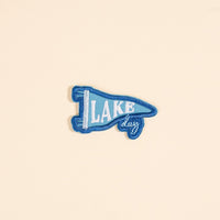 The Darling Effect's Lake Day Mix & Match Iron-On Patch features blue and light blue embroidery with "LAKE" and "day." Perfect for customizing jackets or adding to your patch collection, it's displayed on a plain beige background.