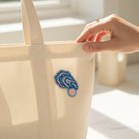 A hand with a ring holds a beige canvas tote bag featuring The Darling Effect's Oyster Mix & Match Iron-On Patch. Add personality to your look with this colorful iron-on design, illuminated by sunlight streaming through the window.