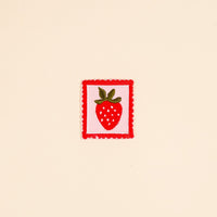 The Darling Effect’s Strawberry Mix & Match Iron-On Patch features a red strawberry with green leaves on a white background and a red scalloped border. Add playful color to your items—shown here on a beige surface.