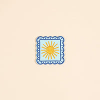 The Darling Effect’s Sunshine Mix & Match Iron-On Patch features a yellow sun with rays on light blue, bordered by blue scallops—a cheerful square accent for your patch collection.