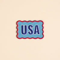 The Darling Effect’s USA Mix & Match Iron-On Patch features bold blue and white “USA” letters with a red wavy border on beige—ideal for customizing jackets or adding to your colorful patch collection.