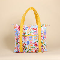 The Darling Effect's Ciao Bella The Everyday Quilted Tote Bag features a vibrant fruit and floral print, wide quilted yellow handles and straps—making it the perfect colorful everyday tote against a beige background.