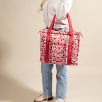 A person in a white top and blue jeans holds The Darling Effect’s Strawberry Fields The Everyday Quilted Tote Bag, which features a large floral pattern, red handles, and ruffle details, standing against a beige background.
