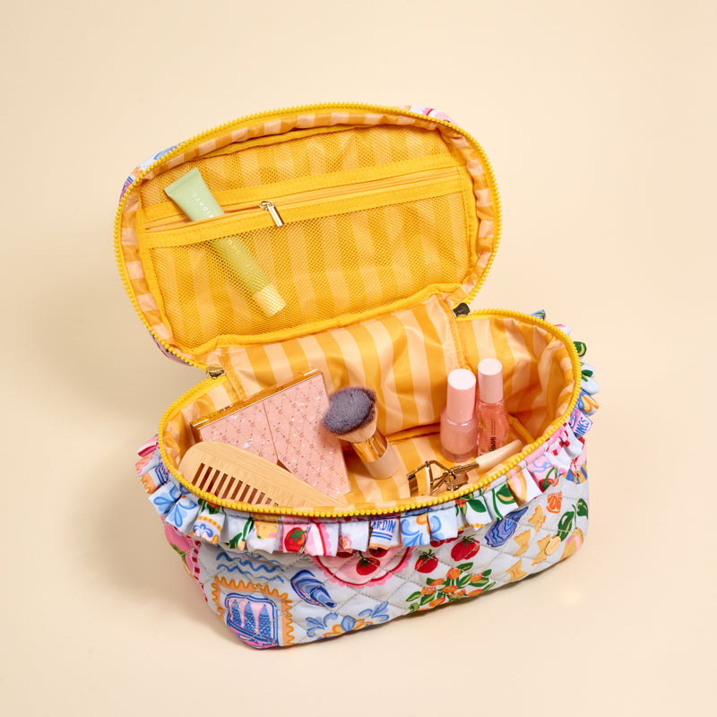 The Darling Effect's Ciao Bella Glam & Go Quilted Cosmetic Bag, featuring a floral pattern and yellow striped interior, is open to reveal cosmetics—a brush, comb, tubes, compact, and a small green tube in the mesh pocket.