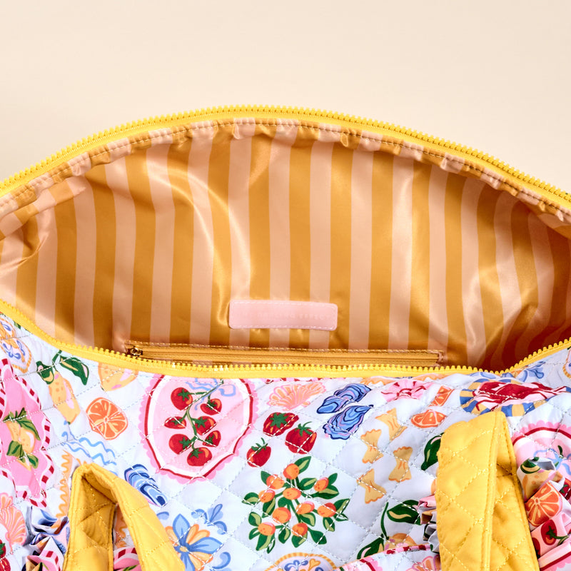 The Darling Effect's Ciao Bella On-the-Go Quilted Weekender Bag features yellow handles and zipper, colorful fruit and floral patterns, a striped interior with zip pocket, and spacious design—combining style and function for your travels.