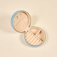 The Darling Effect’s Fruit Market Round Velvet Jewelry Case in light blue is open on a plain surface, showing a beige interior with ring slots, small compartments, and a pocket to organize jewelry.