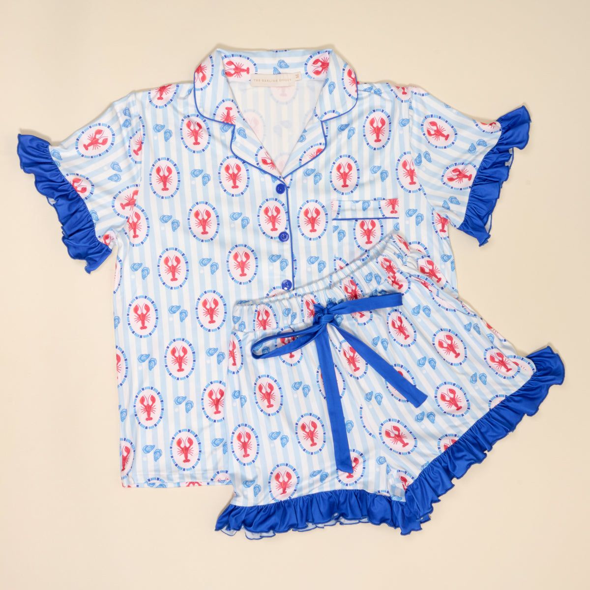 The Darling Effect’s Harbor House Slumber Party Pajama Shorts Set features blue and white stripes, red lobster and anchor patterns, ruffled trim, blue buttons, a ribbon-tie waist, and ultra-soft fabric for cozy comfort.