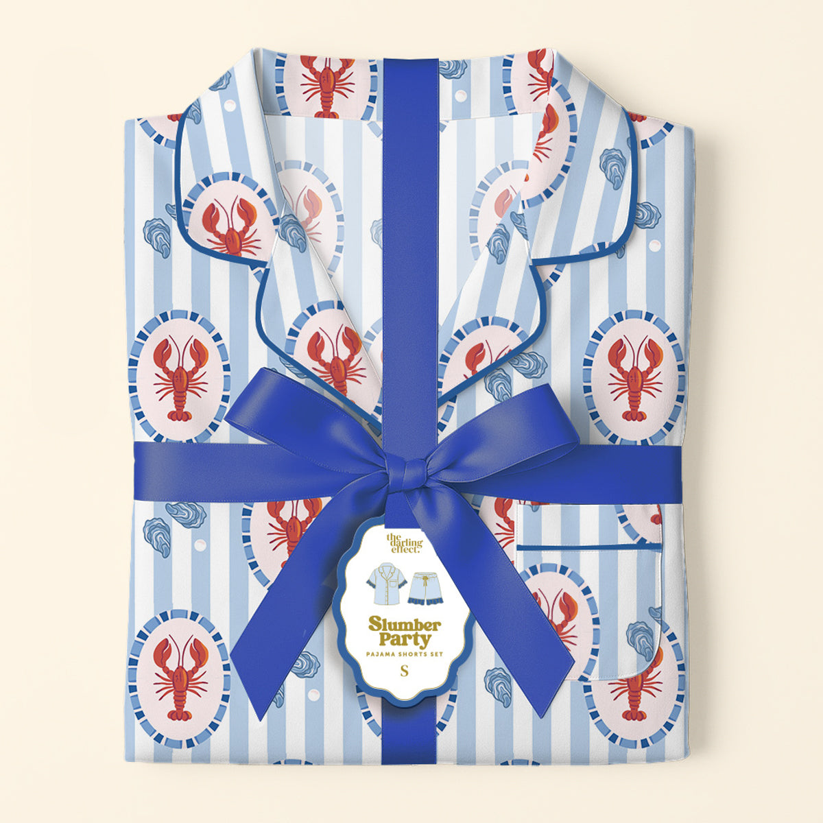 The Darling Effect's Harbor House Slumber Party Pajama Shorts Set, featuring folded blue-and-white stripes with red lobsters, is tied with a ribbon and a tag reading “Slumber Party Pajama Set S,” arranged on a cream background.