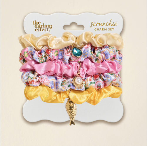 The Darling Effect’s Ciao Bella Scrunchie Charm Set features three vibrant scrunchies—gold, pink, and patterned—each with a unique hair charm: teal gem, pearl, or fish. Stylishly displayed on a branded card, it’s perfect for playful hair accessories.