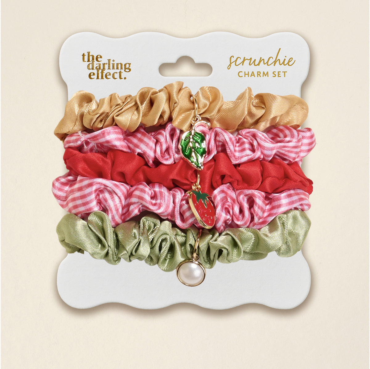 The Darling Effect’s Strawberry Squeeze Scrunchie Charm Set features four stylish hair accessories—gold, red, pink gingham, and sage green—with gold strawberry, leaf, and pearl charms on a white card labeled “scrunchie set.”.