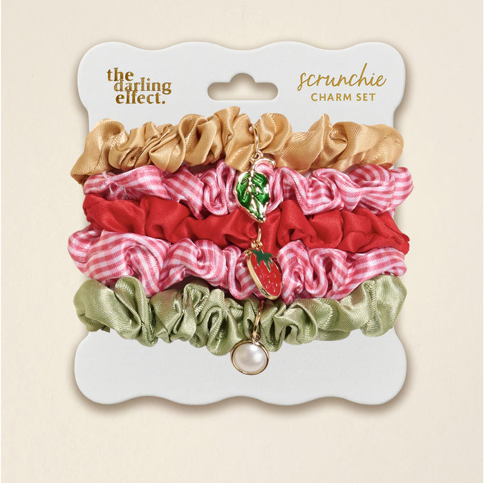 The Darling Effect’s Strawberry Squeeze Scrunchie Charm Set features four stylish hair accessories—gold, red, pink gingham, and sage green—with gold strawberry, leaf, and pearl charms on a white card labeled “scrunchie set.”.