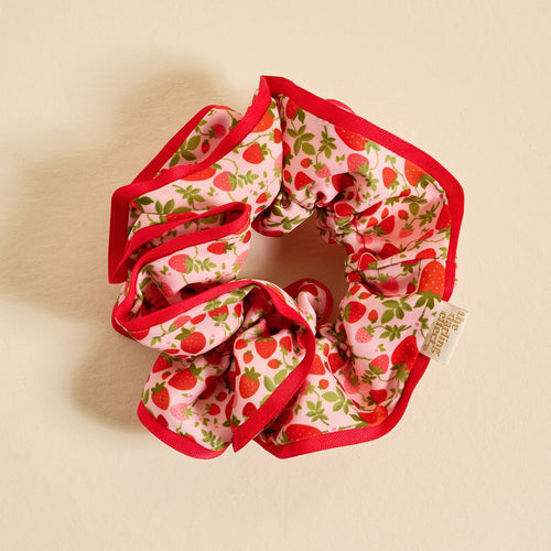The Darling Effect’s Strawberry Fields Ruffle Up Scrunchie features oversized ruffles, a red border, and red strawberries with green leaves on light pink, offering a gentle hold. Shown on a neutral surface.