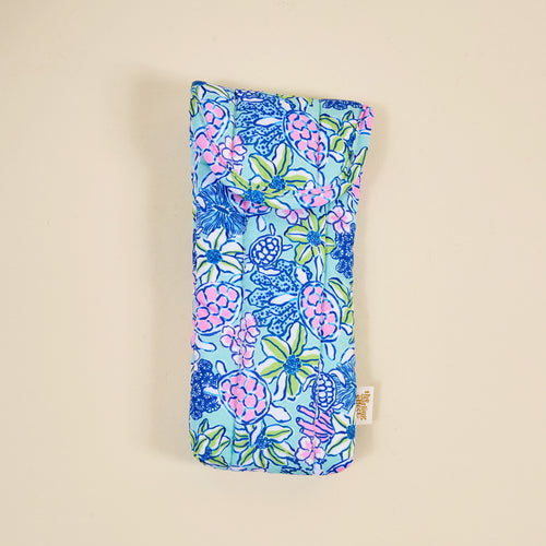 The Coastal Cove Sunglasses Case by the-darling-effect features a soft, quilted design with a blue, pink, and green floral pattern, magnetic flap closure, and a small beige side tag, shown on a plain light background.