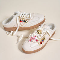 Two white slip-on sneakers with brown soles and fuzzy lining, styled with The Darling Effect's Miss Americana Dazzle Shoe Charm Pack—featuring a gold heart, stars, red bow, flag, and gem charms for a playful look.
