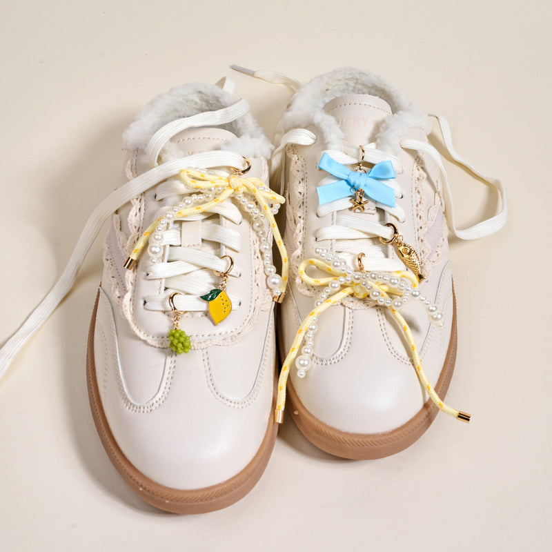 The Darling Effect's Summer Soirée Dazzle Shoe Charm Pack features white sneakers with cream soles, yellow laces, pearls, sneaker charms, pastel blue bows, and a fluffy lining—all shown on a light background.
