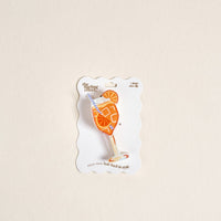 Shaped like an Aperol Spritz complete with a straw, the playful Aperol Spritz Shape Up Claw Clip from The Darling Effect is shown on white packaging featuring the brand name and the phrase “Hold your hair back in style.” The product is set against a plain off-white background.
