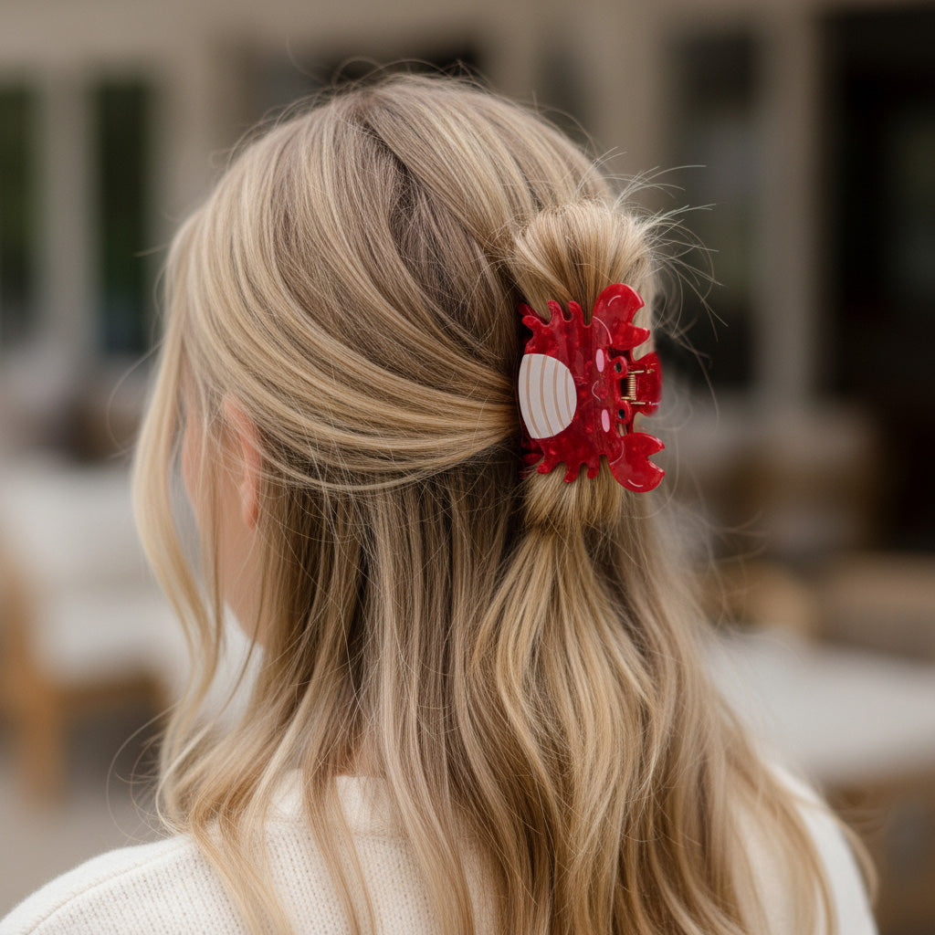 A woman with wavy blonde hair styled in a cute half-up look with The Darling Effect Crab Shape Up Claw Clip (red) and a cream-colored top is seen from behind, the softly blurred background adding to the charm.