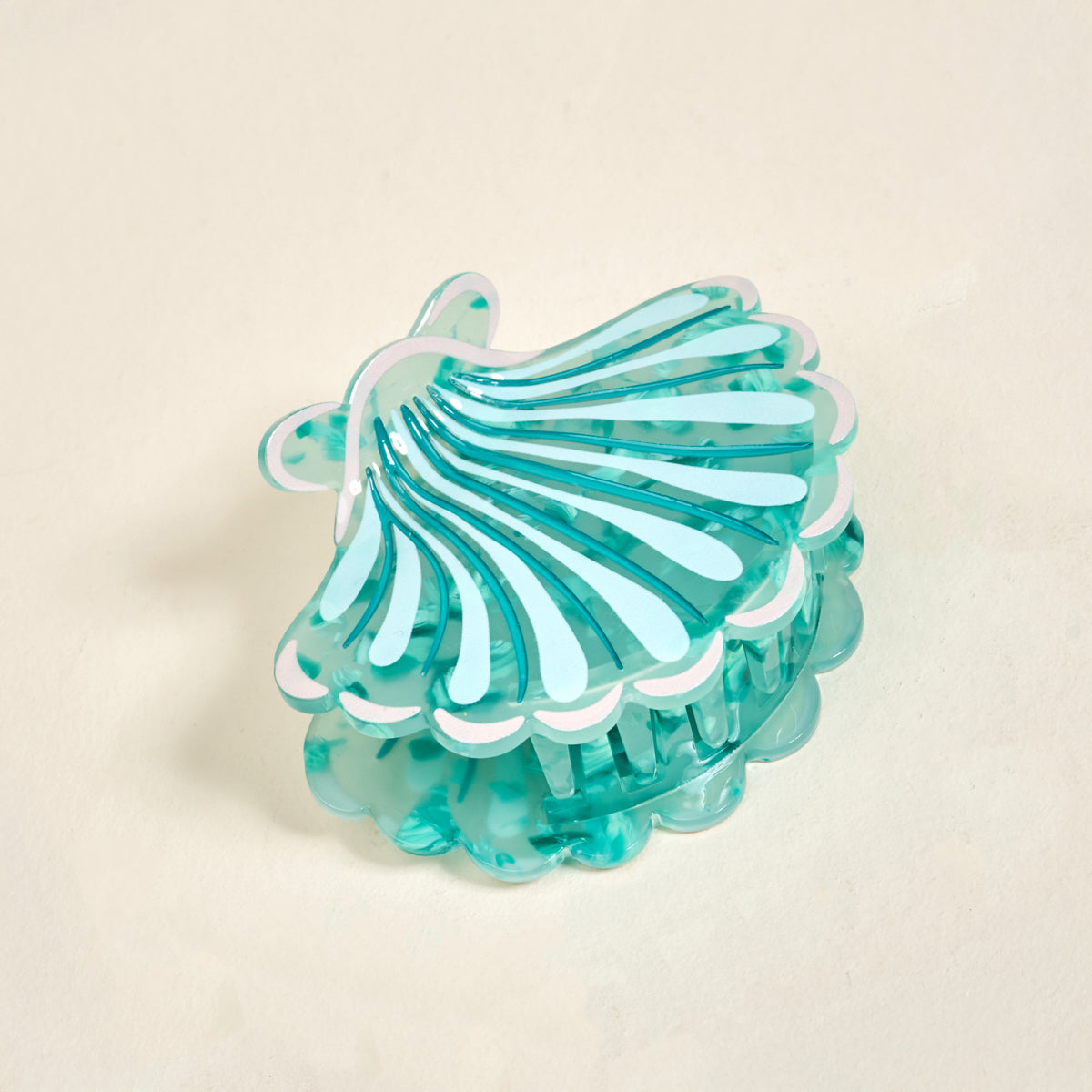 The Darling Effect's Seashell Shape Up Claw Clip features a translucent turquoise seashell design with white edges, making it a charming hair accessory for cute hairstyles. Shown on a light beige surface.