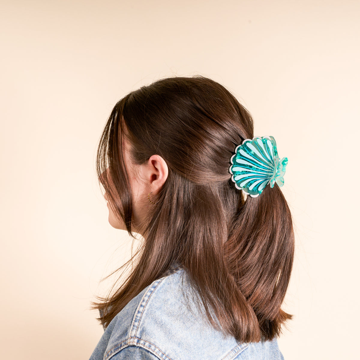 A person with straight brown hair in a light blue denim jacket is shown from the back, wearing The Darling Effect’s Seashell Shape Up Claw Clip—a large, teal shell-shaped accessory securing their cute half-up hairstyle.