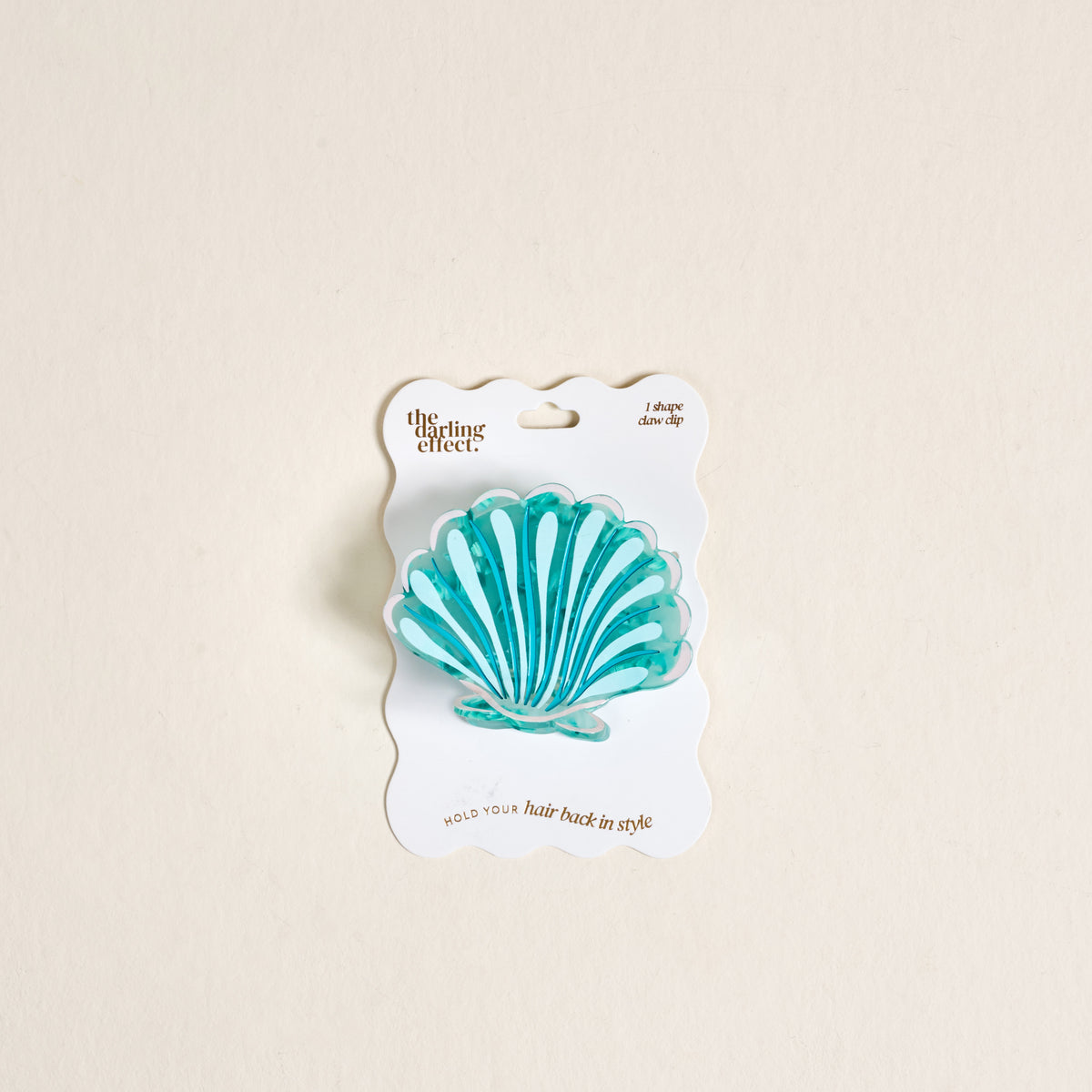 The Seashell Shape Up Claw Clip by The Darling Effect is a blue and turquoise shell-shaped hair accessory on a wavy-edged white card reading “The Darling Effect” and “HOLD your hair back in style for a cute hairstyle.”.