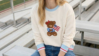Someone wearing The Darling Effect’s Star Spangled Bear Signature Crew—a cream sweatshirt with light blue cuffs and collar, featuring an embroidered bear holding an American flag—sits on metal bleachers.